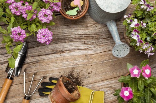 A wooden outdoor table in a garden setting, featuring several potted flowering plants with pink, purple, and white blooms arranged along the edges. The table surface shows visible wood grain and overall weathered texture. Gardening tools, including a small hand rake, a trowel, and a gardening fork with wooden handles, are placed on the table, along with a pair of yellow gardening gloves. A small terracotta pot with soil and a green plant, as well as decorative garden ornaments with pastel-colored shapes, are also visible. In the background, neighboring potted plants and garden equipment suggest an actively maintained outdoor space, potentially part of a back garden in Barnet. The scene is lit with natural daylight, indicating a clear, bright day suitable for gardening activities, aligning with services offered by Landscapers Barnet for garden and outdoor space improvements.