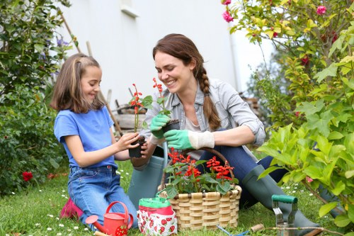 A woman and a young girl are gardening together in a lush, well-maintained backyard garden that features a variety of plants, shrubs, and flowering bushes, with green grass covering the ground. The woman, wearing a light grey shirt and green gardening gloves, is smiling as she hands a potted plant with red flowers to the girl, who is dressed in a blue t-shirt and jeans. They are sitting on the grass amidst gardening tools and a wicker basket filled with gardening supplies, including a small watering can with a floral pattern. The garden background is filled with leafy green bushes and colorful flowering plants, suggesting a vibrant outdoor space typical for gardening activities in Barnet, UK. The overall scene is well-lit with natural daylight, indicating clear weather, and conveys a peaceful, productive outdoor environment suitable for gardening and landscaping services offered by Landscapers Barnet.