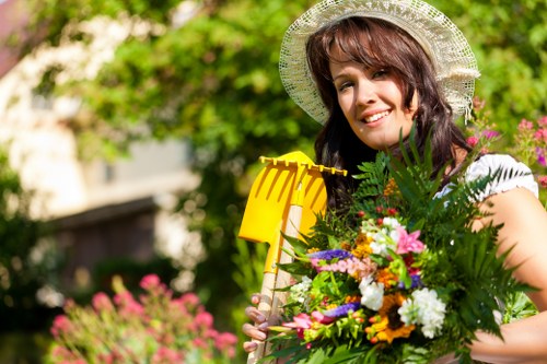 A woman with shoulder-length brown hair wearing a wide-brimmed straw hat is standing in a lush garden during daytime. She is smiling and holding a yellow garden rake in her right hand, while her left arm is wrapped around a vibrant bouquet of mixed flowers and green foliage. The background features a well-maintained lawn with dense greenery, including bushes and trees, along with colorful flowering plants and blurred pink flowers in the distance. The scene is illuminated by natural daylight, indicating a bright, sunny day, and the garden appears to be part of a residential outdoor space, suitable for gardening and landscaping activities with the assistance of professional services from Landscapers Barnet.
