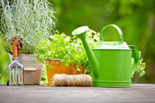 A close-up view of a garden table surface featuring various gardening tools and supplies, including a bright green metal watering can, a coiled natural jute plant tie, and several potted herbs and plants with green foliage. The background shows a lush, out-of-focus green garden with trees and shrubs, indicating a well-maintained outdoor space typical of a suburban garden in Barnet. The scene highlights the tools and elements used in gardening and outdoormaintenance, suitable for landscaping services offered by Landscapers Barnet, with a focus on garden care and plant cultivation, under natural daylight conditions.